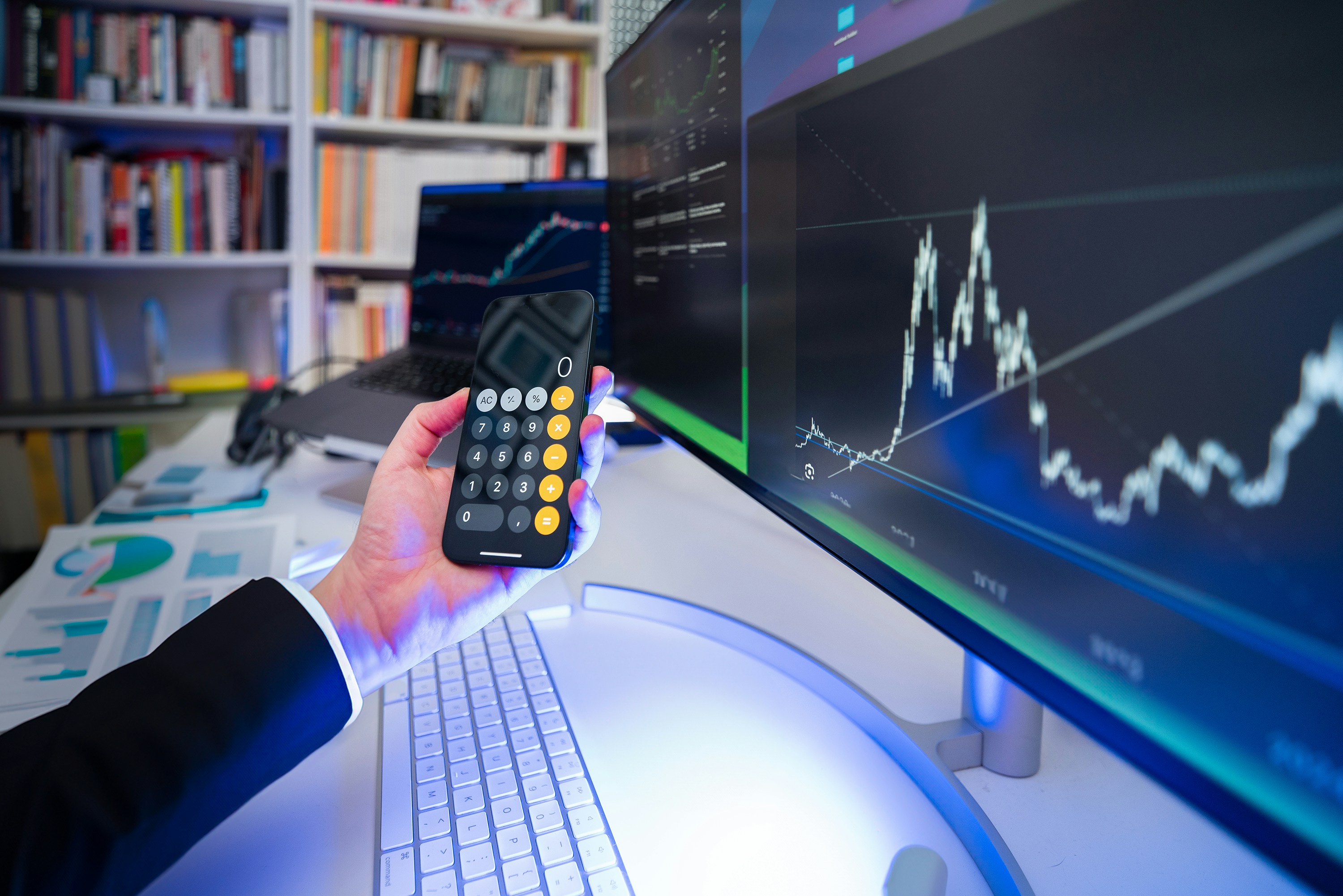 Hand holding a calculator in front of large monitors displaying financial graphs and data.