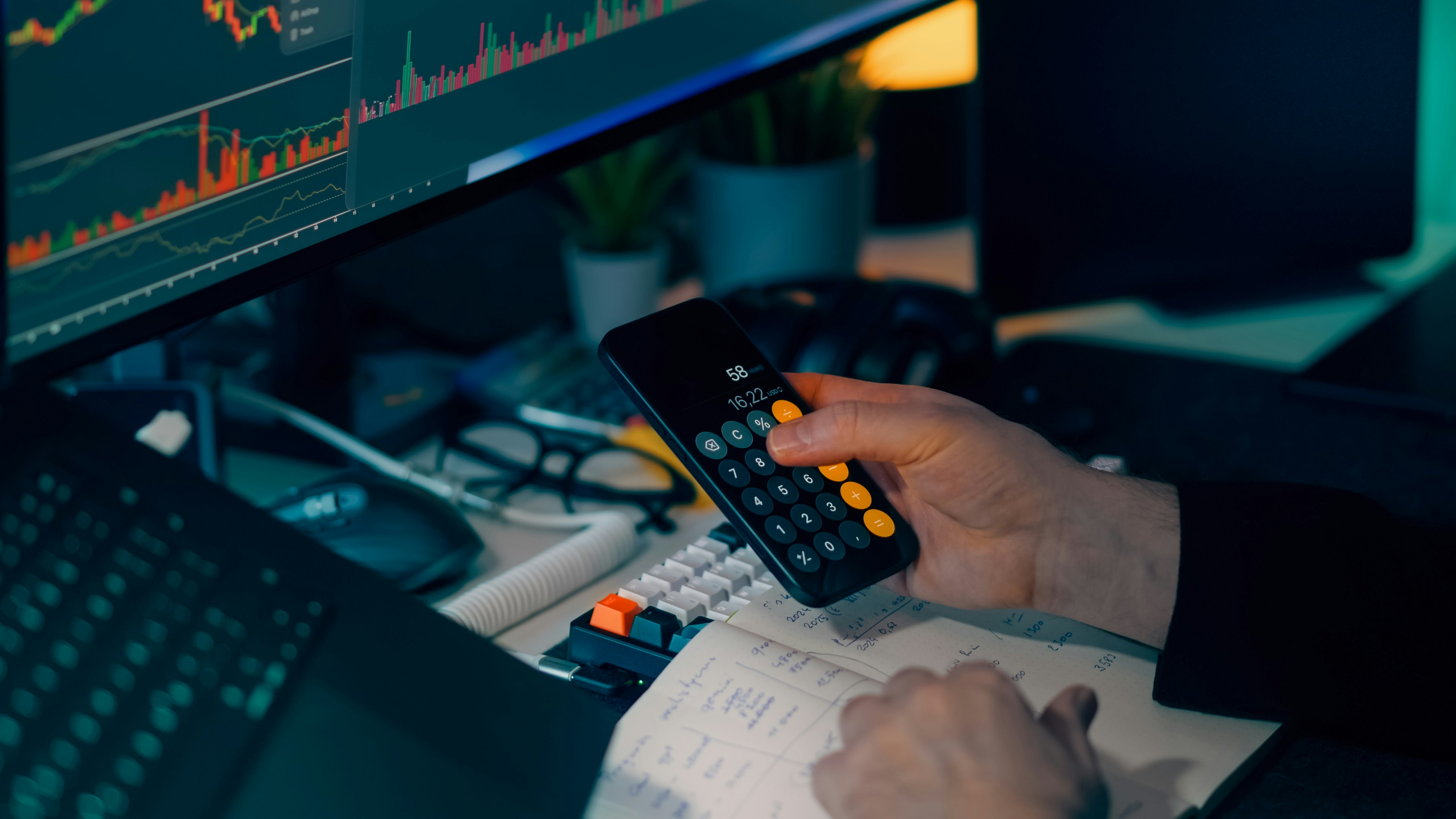 Person holding a calculator over a notebook while financial charts are displayed on a computer monitor