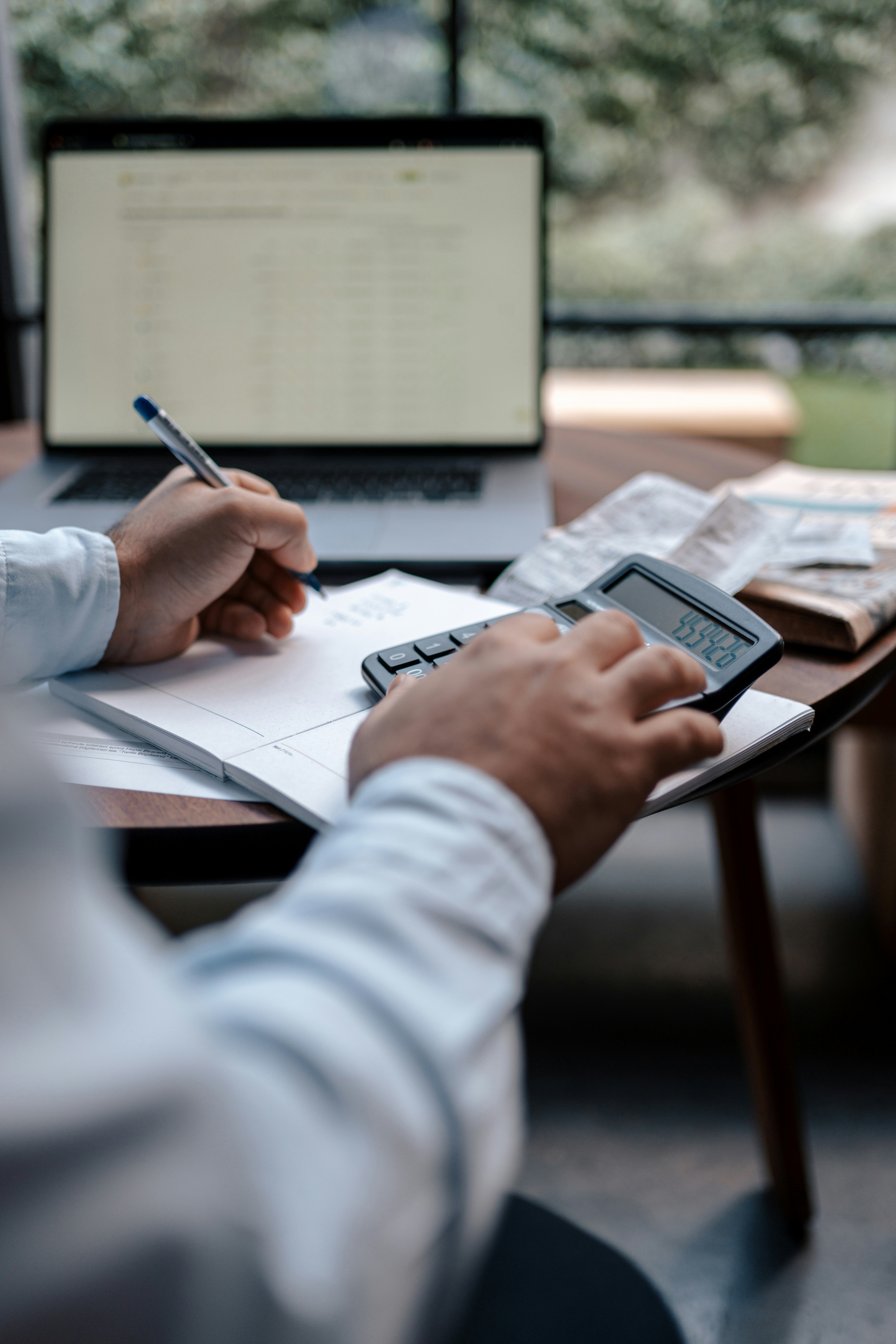 Close-up of a person calculating numbers in a notebook beside an open laptop and receipts.