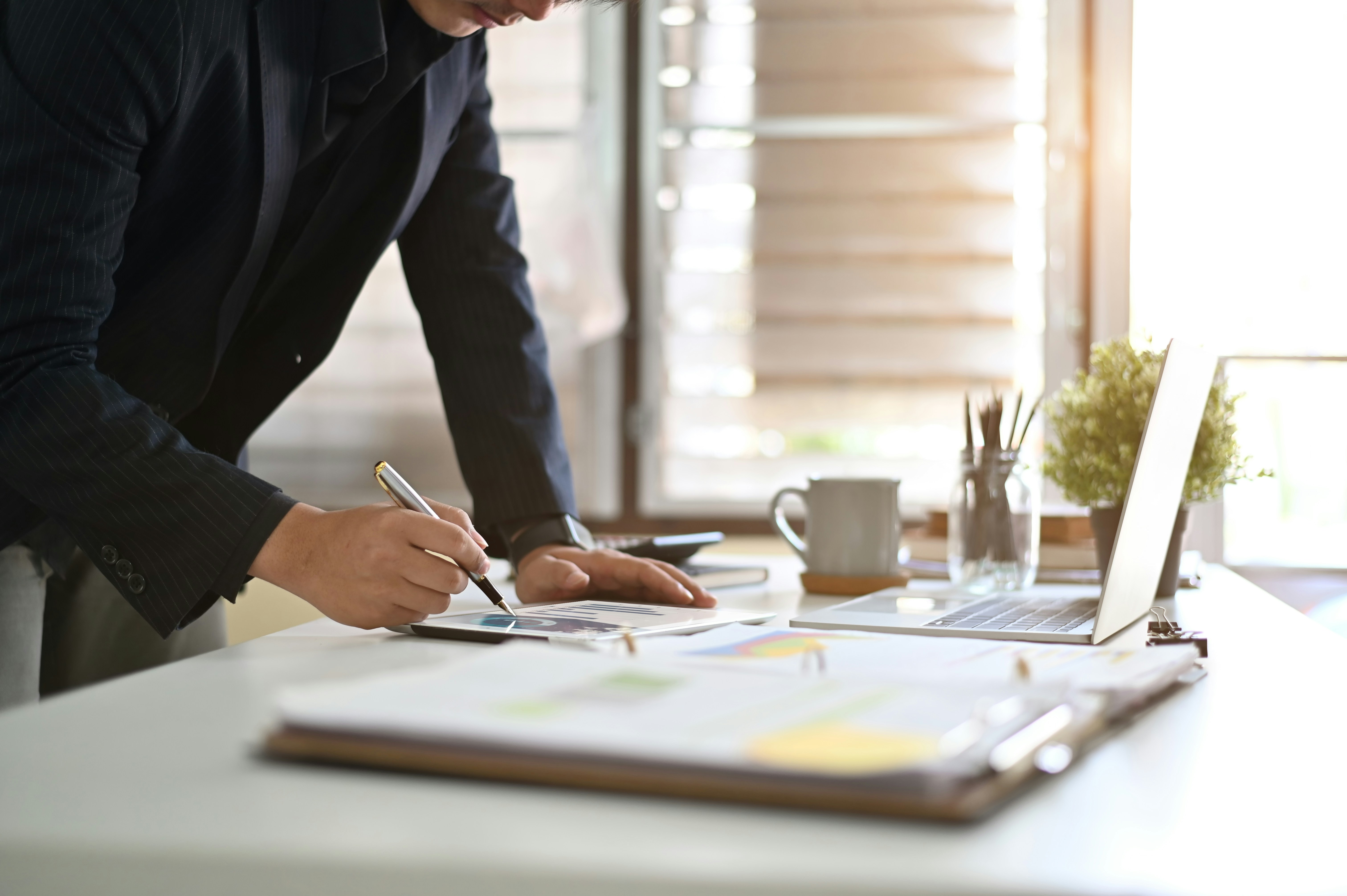 Close-up of a person reviewing papers and writing notes at a desk with a laptop nearby.