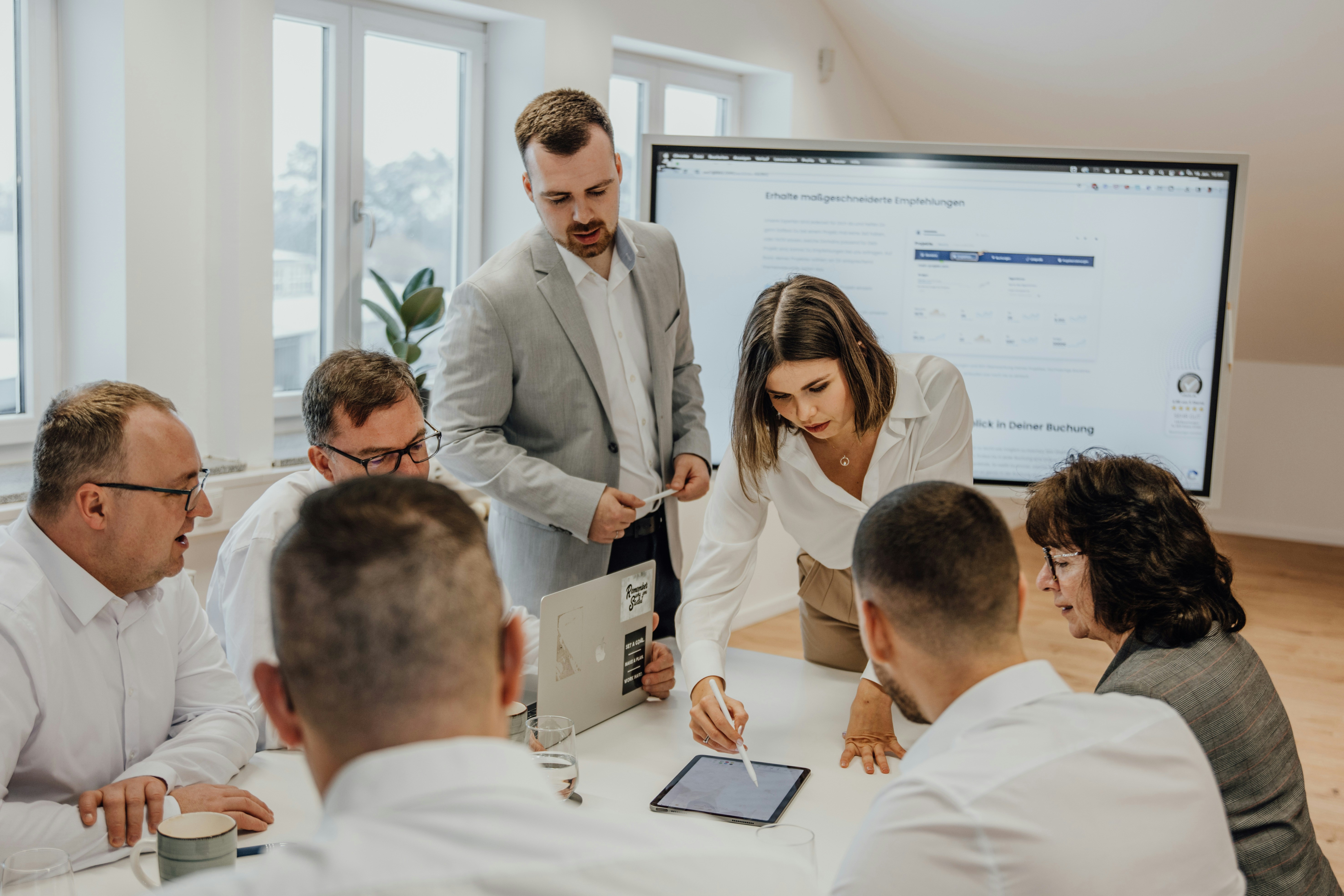 Group of people gathered around a table while one person points to a tablet during a meeting.